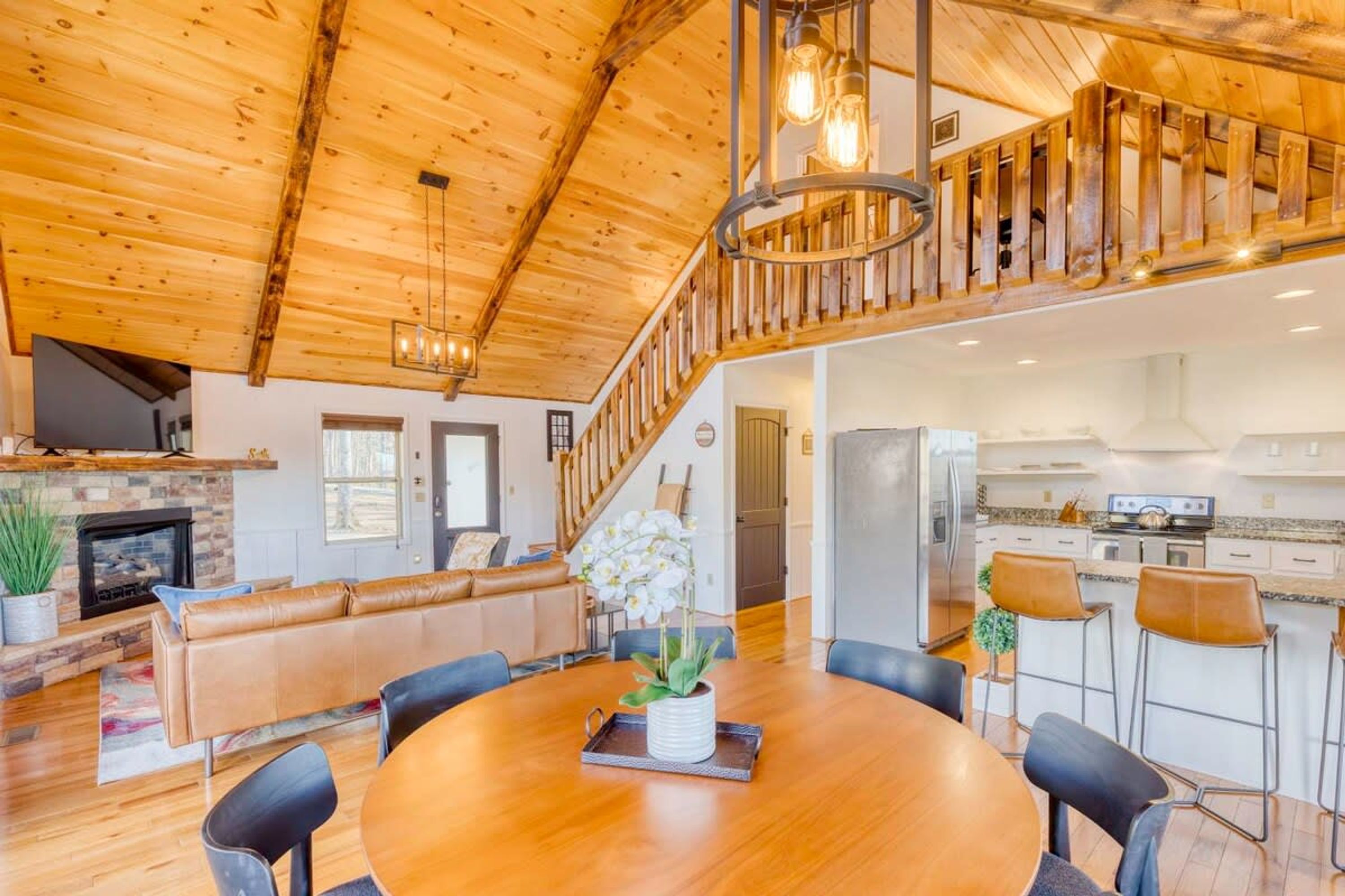 A spacious living area with a wooden ceiling, a fireplace, a dining table with chairs, and a kitchen area.