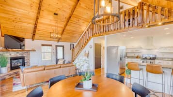 A spacious living area with a wooden ceiling, a fireplace, a dining table with chairs, and a kitchen area.