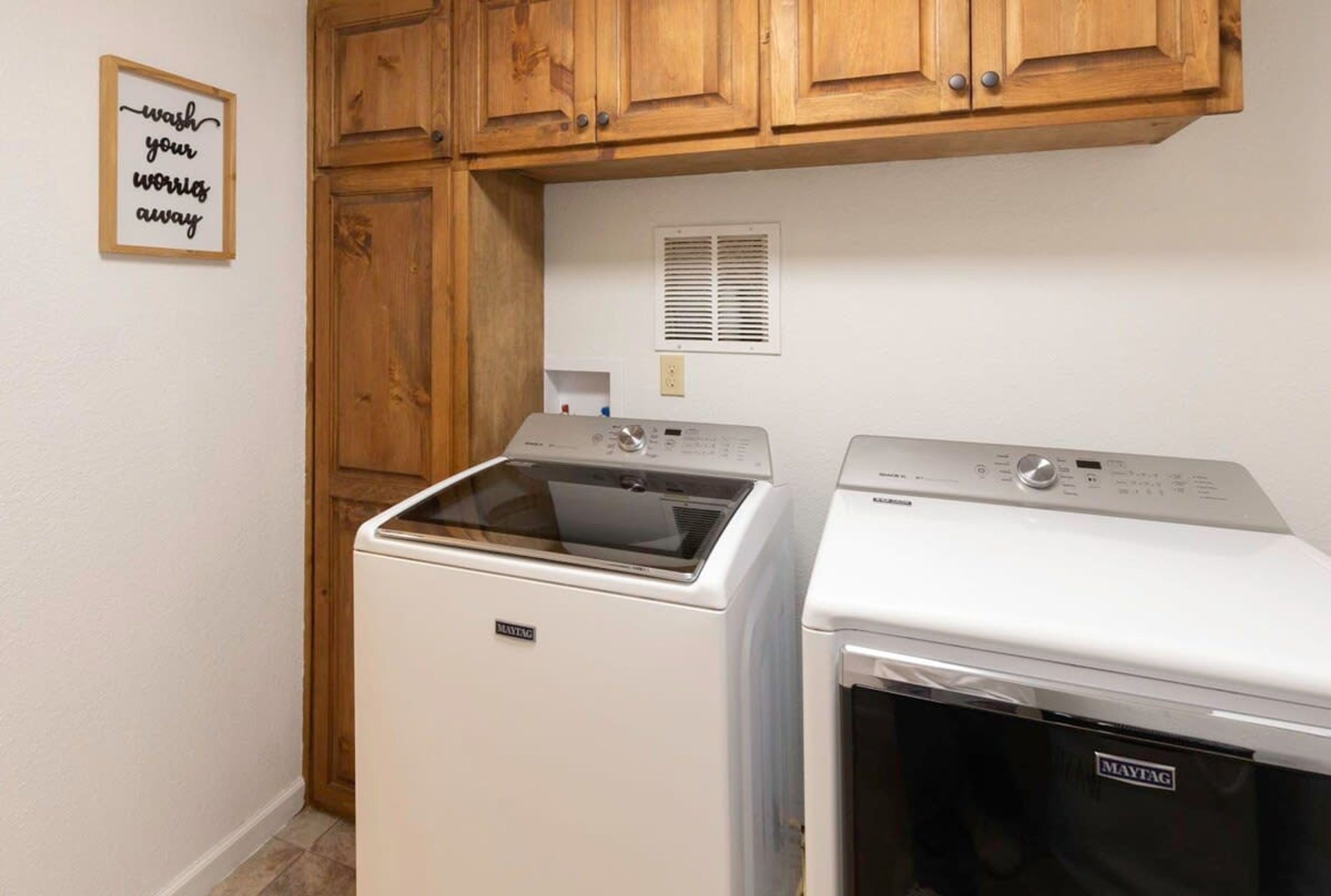 Uma lavanderia com máquina de lavar e secadora Maytag, armários de madeira, um sistema de ventilação e uma frase motivacional na parede.