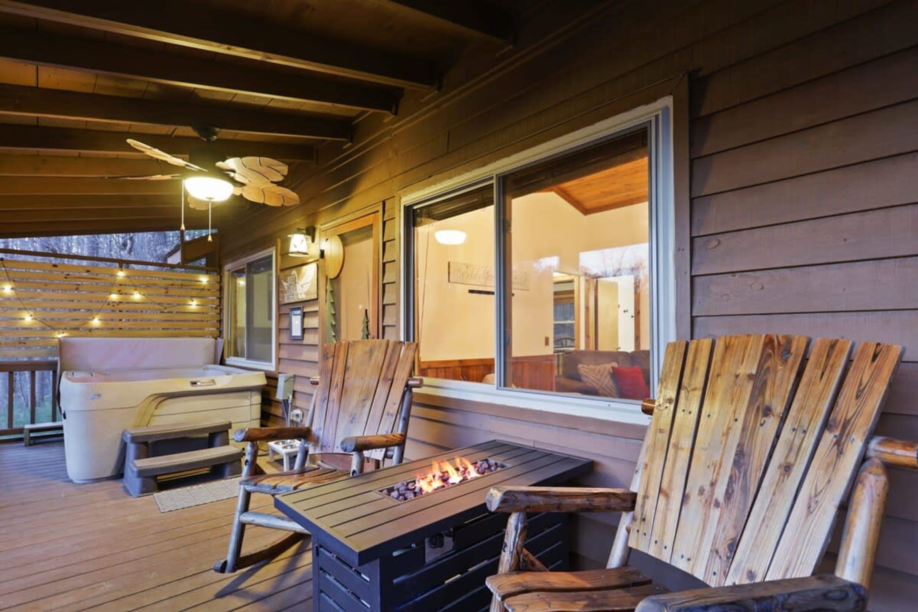 Une terrasse en bois avec des meubles d’extérieur, un foyer et une grande fenêtre donnant sur l’intérieur d’un chalet.