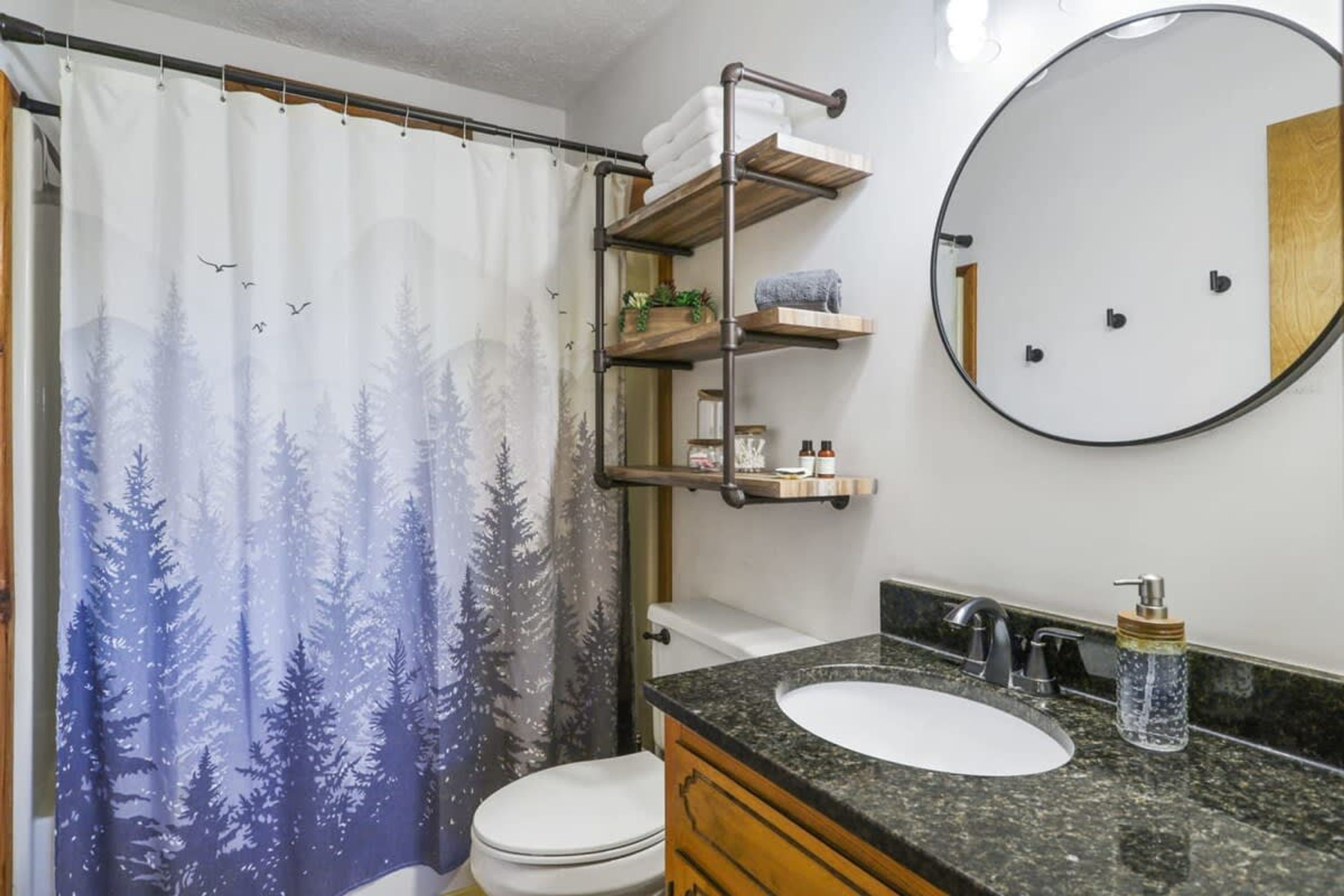 Ein Badezimmer mit Duschvorhang mit Waldmotiv, einer Toilette, einem Waschbecken mit Marmorplatte und einem Spiegel.
