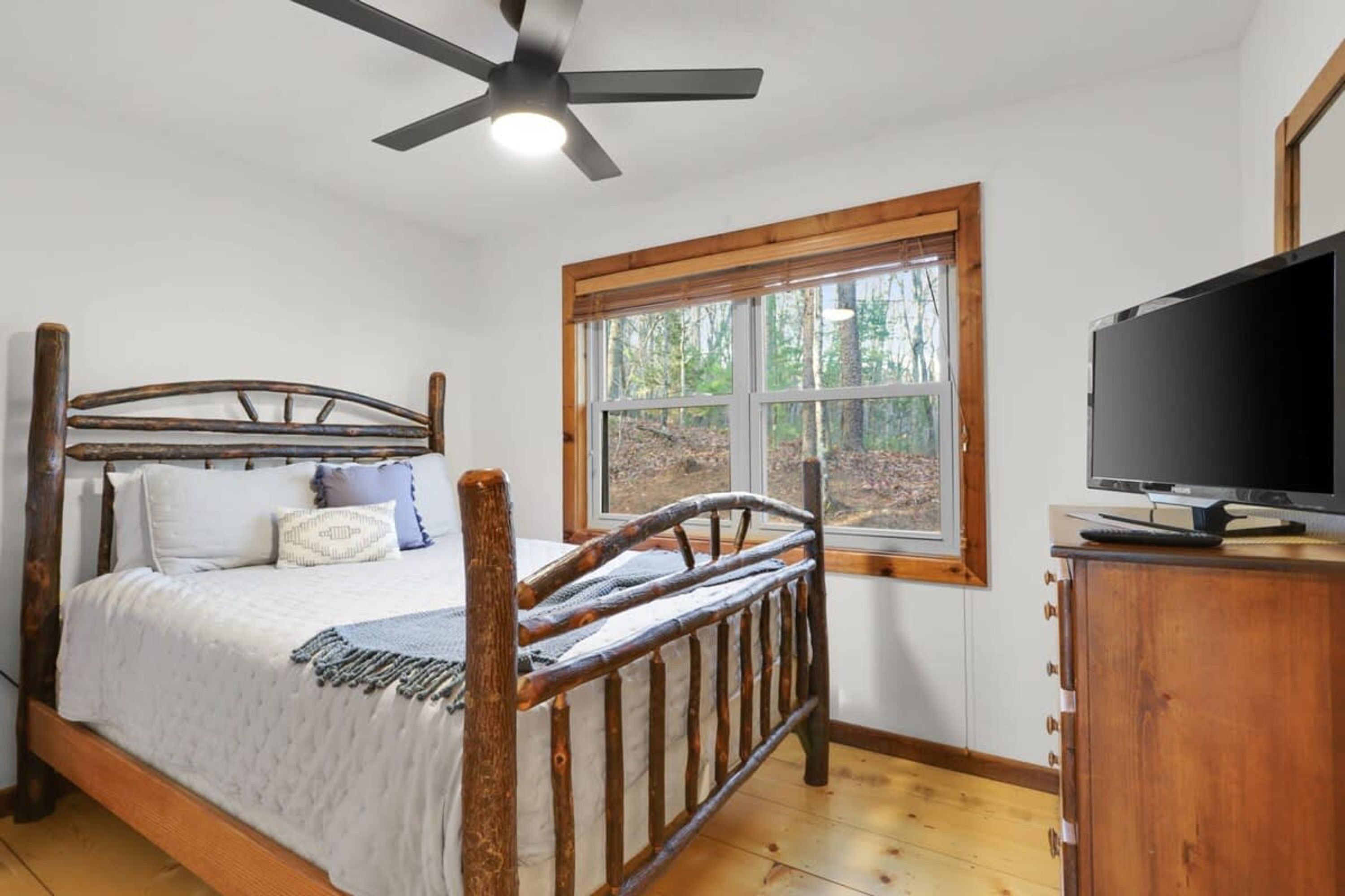 Un dormitorio con cama de madera, ventilador de techo, ventana con vista a árboles y televisor sobre un mueble de madera.