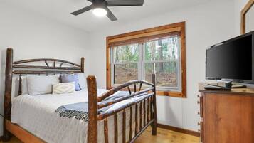 Un dormitorio con cama de madera, ventilador de techo, ventana con vista a árboles y televisor sobre un mueble de madera.