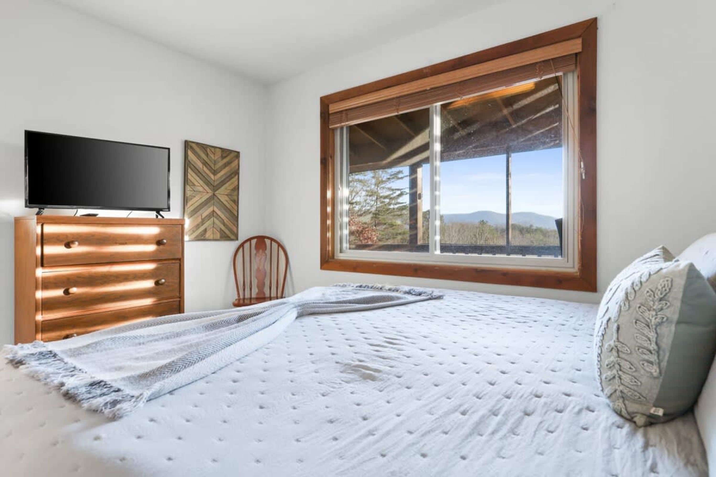 A bedroom with a bed, a dresser, a television, and a window with a view of the outdoors.
