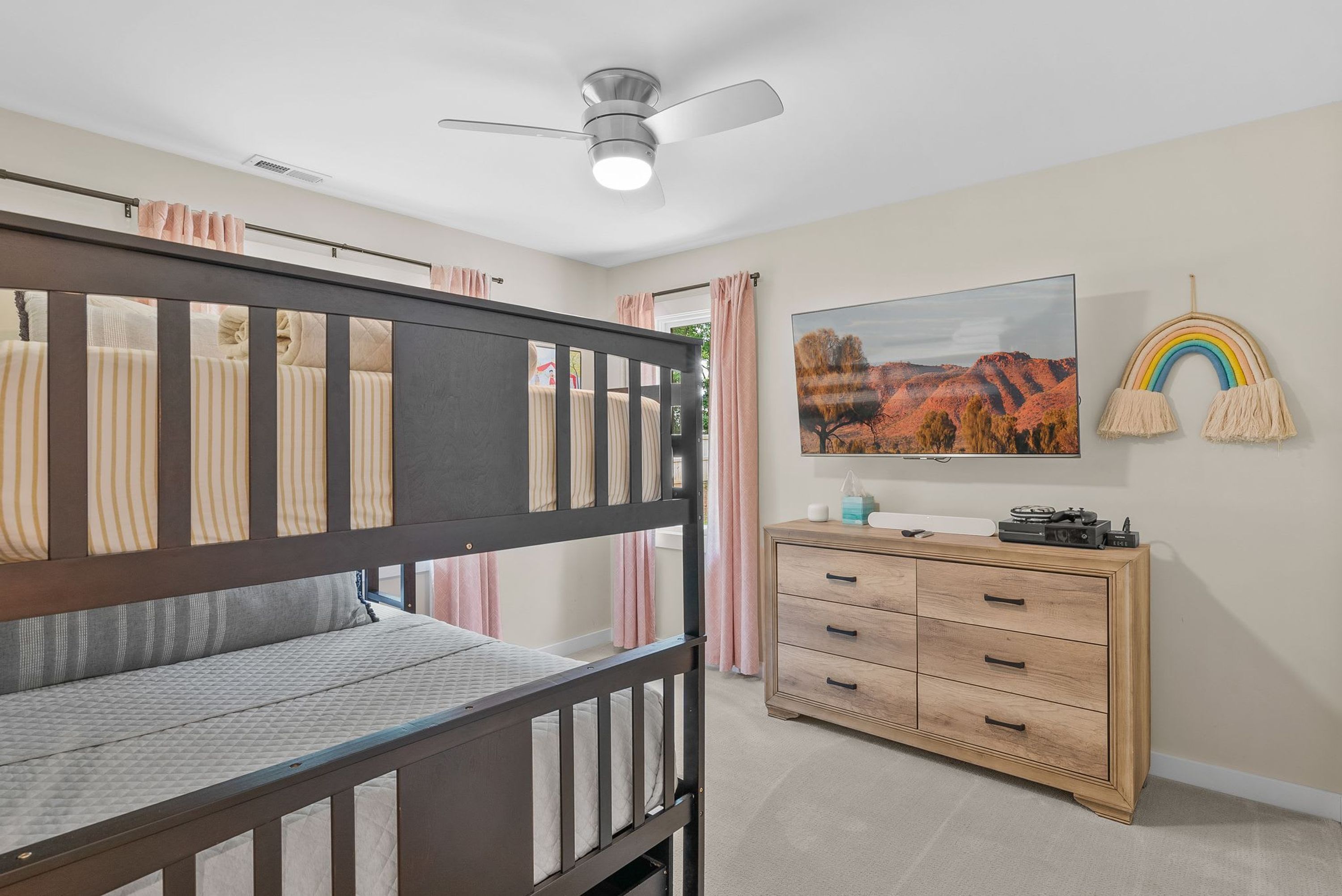 A child's bunk bed with a ceiling fan, a dresser with drawers, a television, and a wall-mounted rainbow decoration.