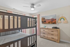 Ein Kinderstockbett mit Deckenventilator, eine Kommode mit Schubladen, ein Fernseher und eine Wanddekoration mit Regenbogen-Motiv.