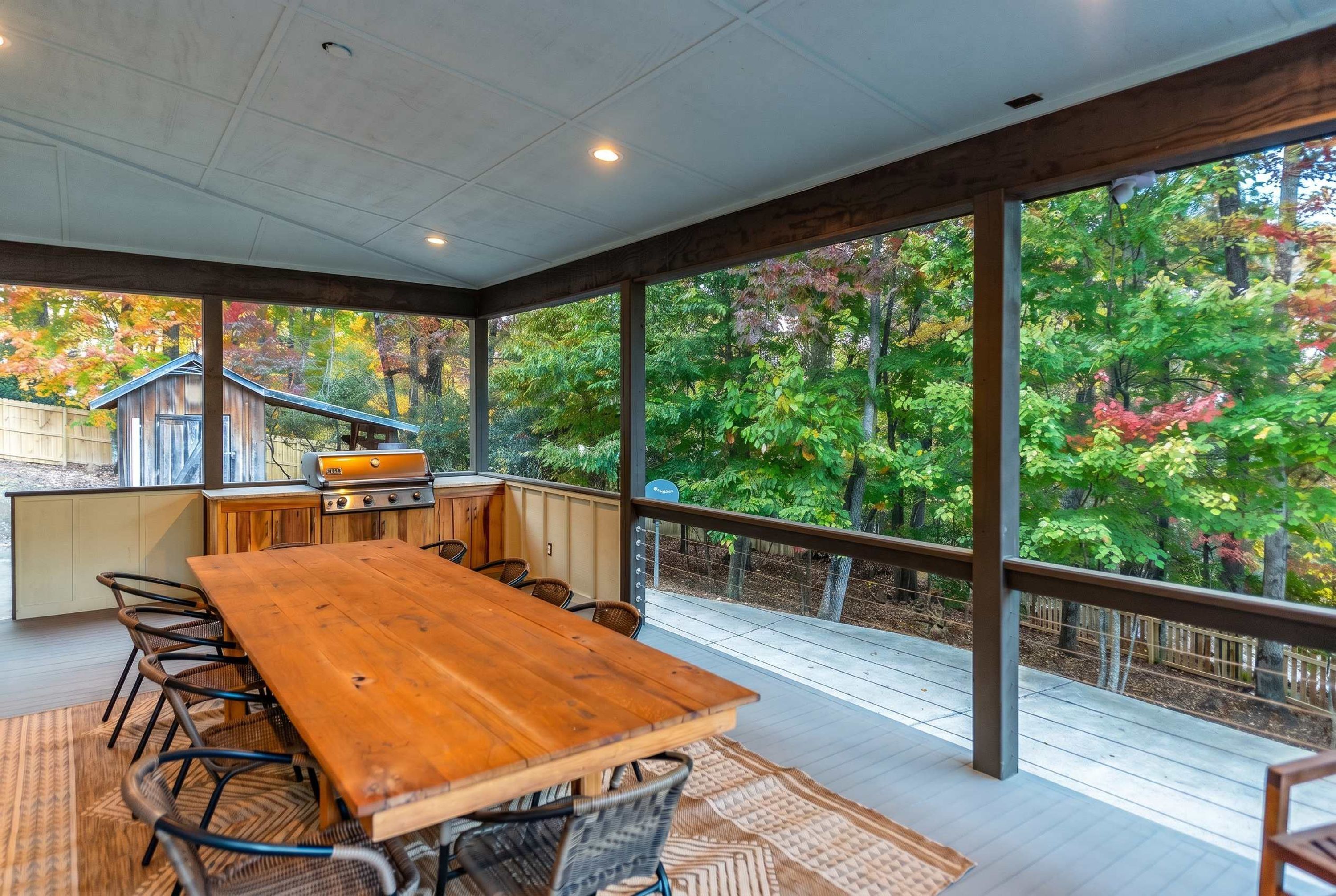 A covered outdoor dining area with a wooden table and chairs, a grill, and a view of a wooded area.