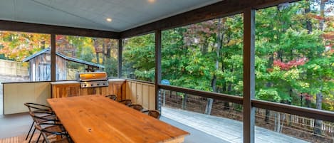 A covered outdoor dining area with a wooden table and chairs, a grill, and a view of a wooded area.