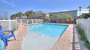 A rectangular swimming pool with a white deck, surrounded by a wooden fence, and blue chairs along the edge. - Toucan Tango- Golf Cart Friendly 3BD Home (Port Aransas)