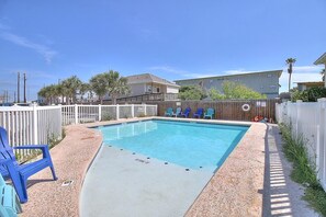 A rectangular swimming pool with a white deck, surrounded by a wooden fence, and blue chairs along the edge. - Toucan Tango- Golf Cart Friendly 3BD Home (Port Aransas)
