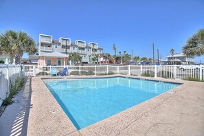 A rectangular swimming pool with a white fence surrounding it, set against a backdrop of multi-story buildings and palm trees. - Toucan Tango- Golf Cart Friendly 3BD Home (Port Aransas)