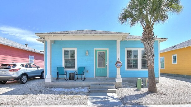 A two-story house with a blue exterior, a front porch with two chairs, and a palm tree. - Toucan Tango- Golf Cart Friendly 3BD Home (Port Aransas)