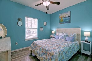 A bedroom with a bed, two bedside tables, a ceiling fan, a window with blinds, and a painting on the wall. - Toucan Tango- Golf Cart Friendly 3BD Home (Port Aransas)