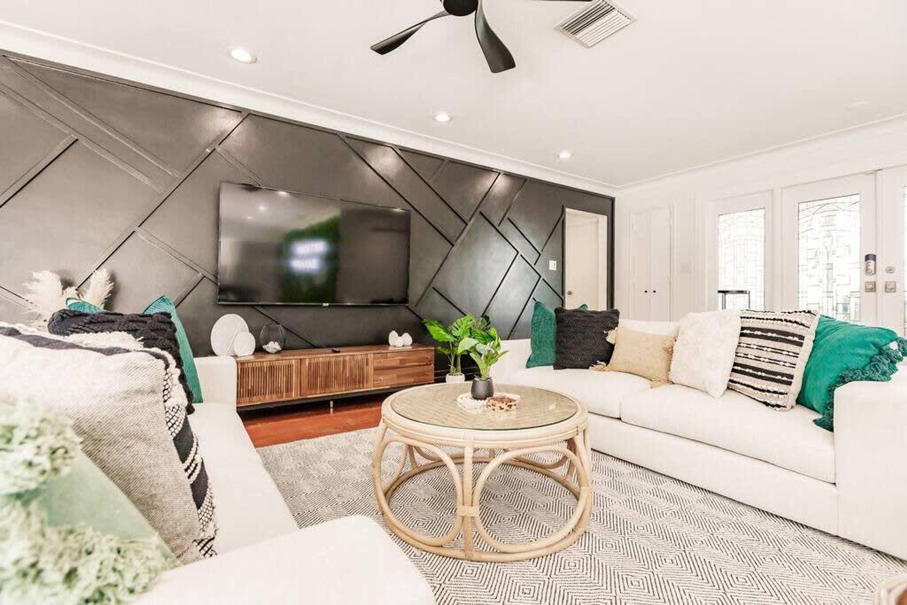 Un salon moderne avec un téléviseur à écran plat fixé sur un mur orné d’un motif géométrique, un canapé blanc agrémenté de coussins verts et à rayures, une table basse ronde en osier et un ventilateur de plafond.