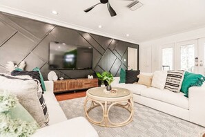 Un salon moderne avec un téléviseur à écran plat fixé sur un mur orné d’un motif géométrique, un canapé blanc agrémenté de coussins verts et à rayures, une table basse ronde en osier et un ventilateur de plafond.