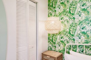 Una habitación con un armario blanco, una cama con ropa de cama blanca, una mesita de noche de madera y una pared con un estampado de hojas tropicales.