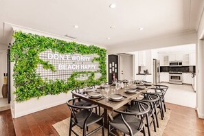 A modern dining area with a long table set for six, surrounded by chairs, and a wall with a green plant wall and motivational text. - Resort Style Backyard Heated Pool Hot Tub Fire Pit (Fort Lauderdale)