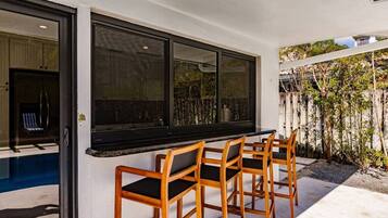 Un patio con piscina, sillas de madera y un amplio ventanal con vistas a un jardín.