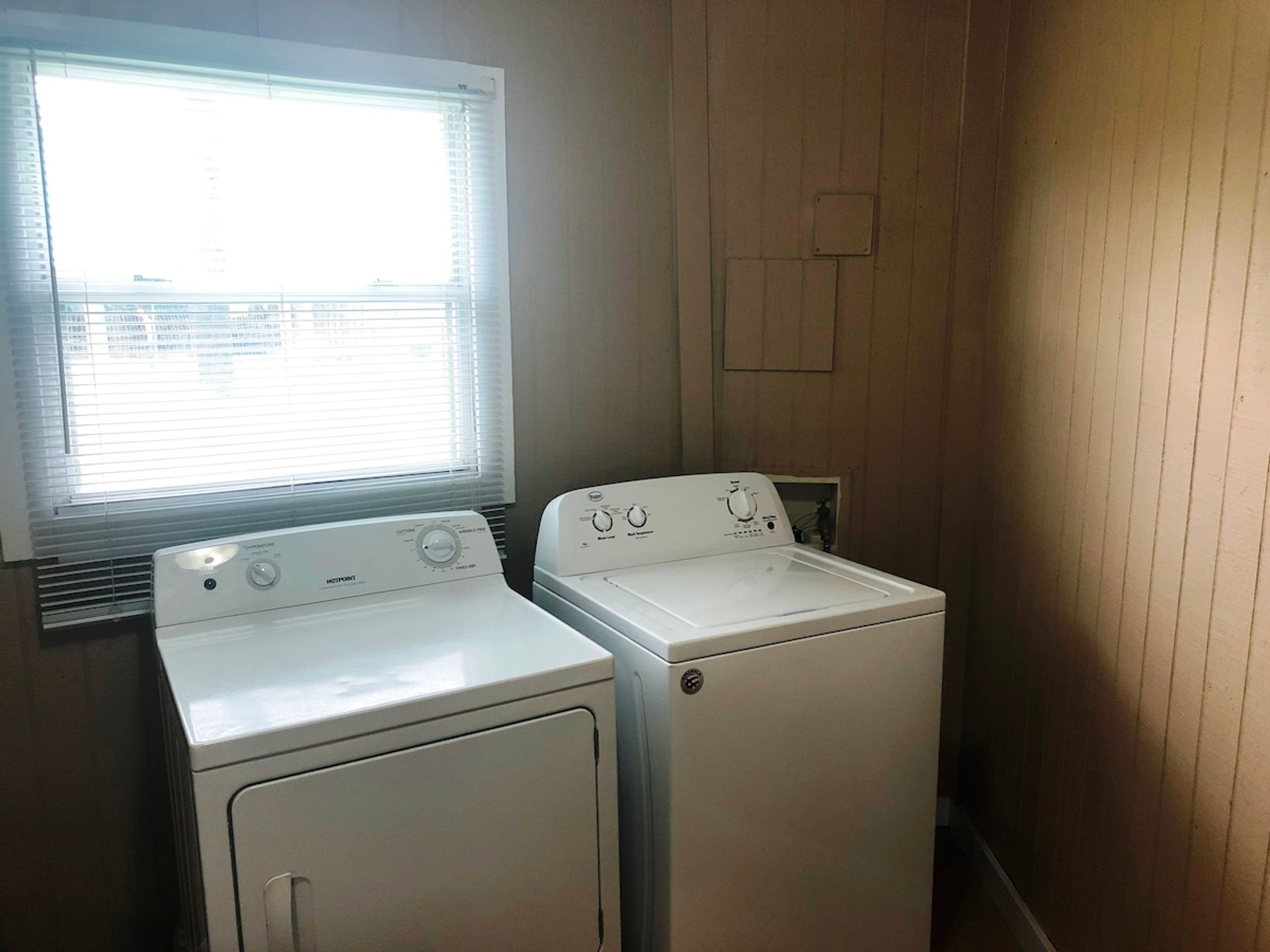 A laundry room with two white washing machines and a window with blinds.