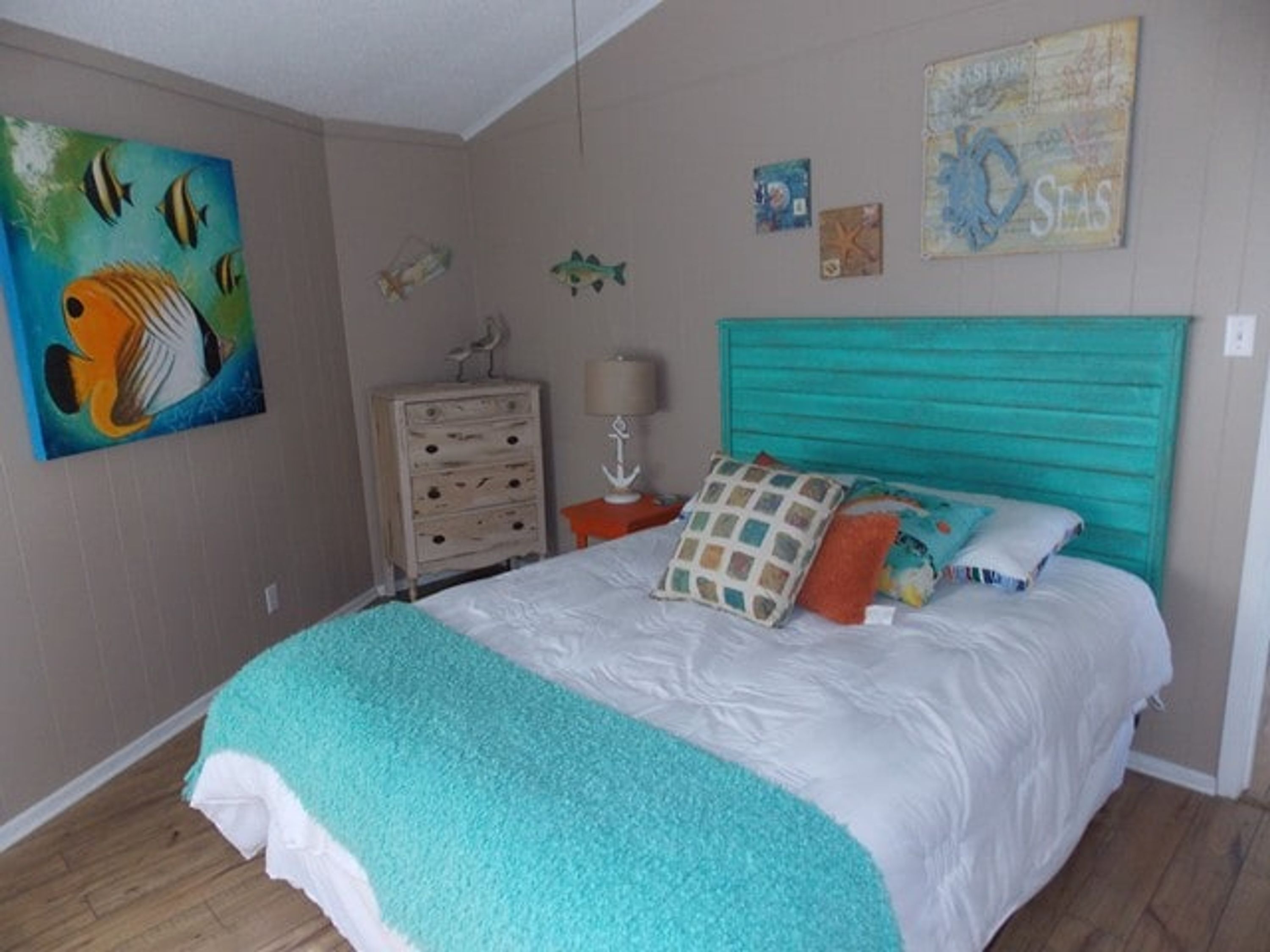 A bedroom with a bed, a dresser, a lamp, and wall decorations including a fish painting and a seashell map.