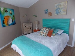 A bedroom with a bed, a dresser, a lamp, and wall decorations including a fish painting and a seashell map. - Beach Shak- Pool View Private Apartment (Port Aransas)