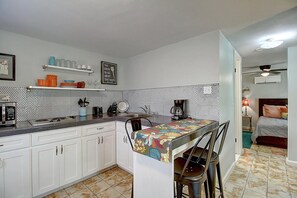 A compact kitchen with white cabinets, a tiled backsplash, and a small dining area with a colorful tablecloth. - Kona Cottage Dog Friendly In Town Location (Port Aransas)