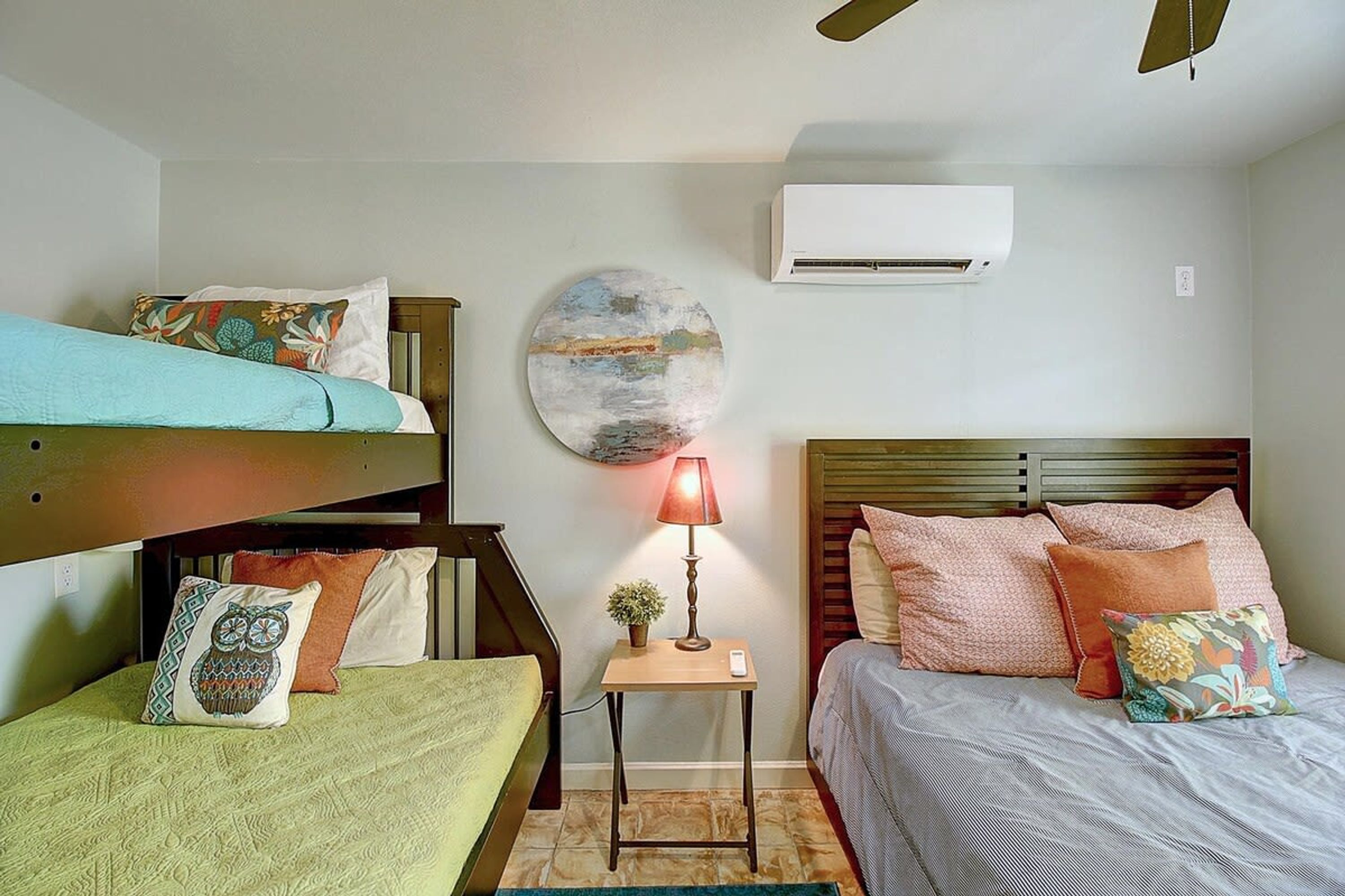 Habitación tipo dormitorio con literas, una mesita de noche con lámpara, aire acondicionado de pared y un cuadro redondo.