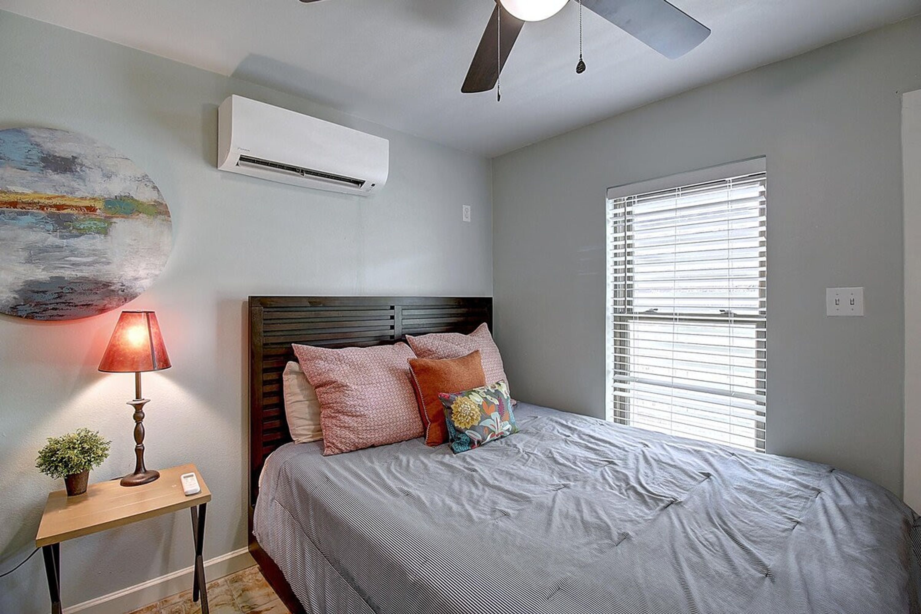 Un dormitorio con cama, mesita de noche, lámpara, ventilador de techo y ventana con persianas.