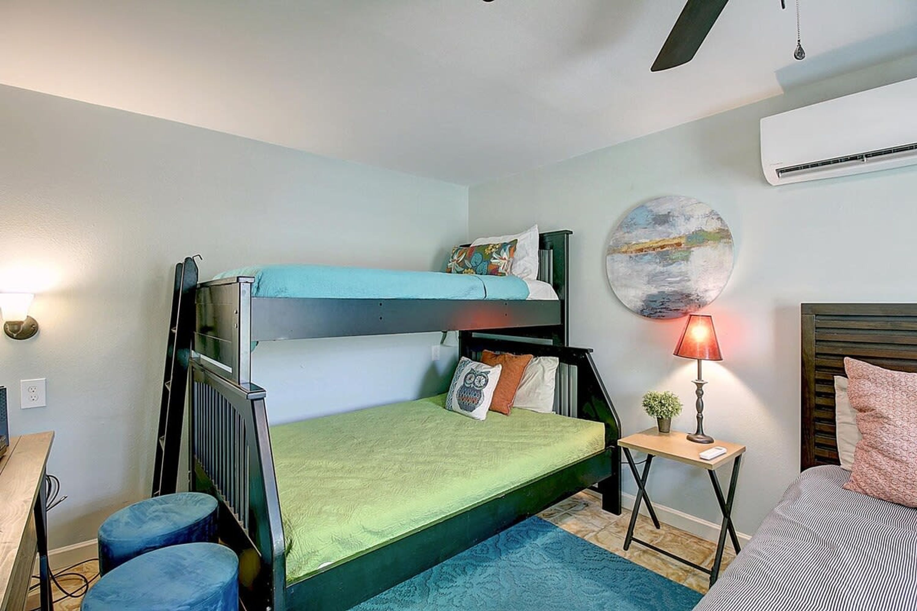 Uma cama de beliche com colchão verde, uma mesinha de cabeceira com abajur e um quadro redondo na parede.