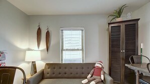A living room with a sofa, a lamp, a window with blinds, a cabinet, and a small table. - Kona Cottage Dog Friendly In Town Location (Port Aransas)