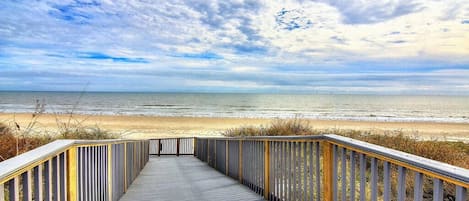 Une promenade en bois qui mène à une plage, d’où l’on a une vue dégagée sur l’océan et sur un ciel partiellement nuageux.