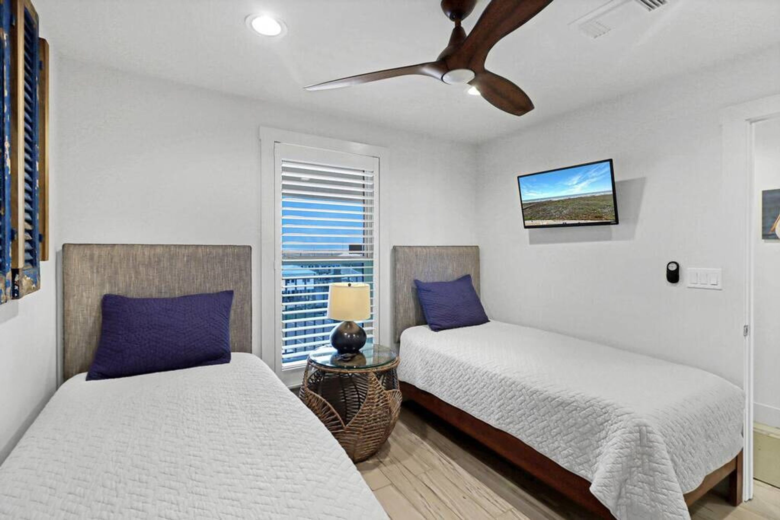 A bedroom with two beds, a ceiling fan, a window with blinds, a bedside lamp, and a wall-mounted picture.