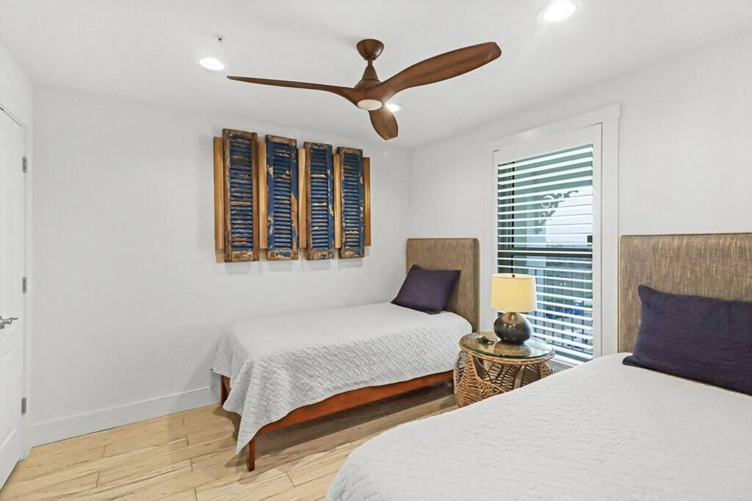 A bedroom with two beds, a ceiling fan, a window with blinds, and a small table with a lamp.