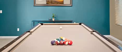 A pool table with balls, a framed picture on the wall, and a potted plant on a blue console table.