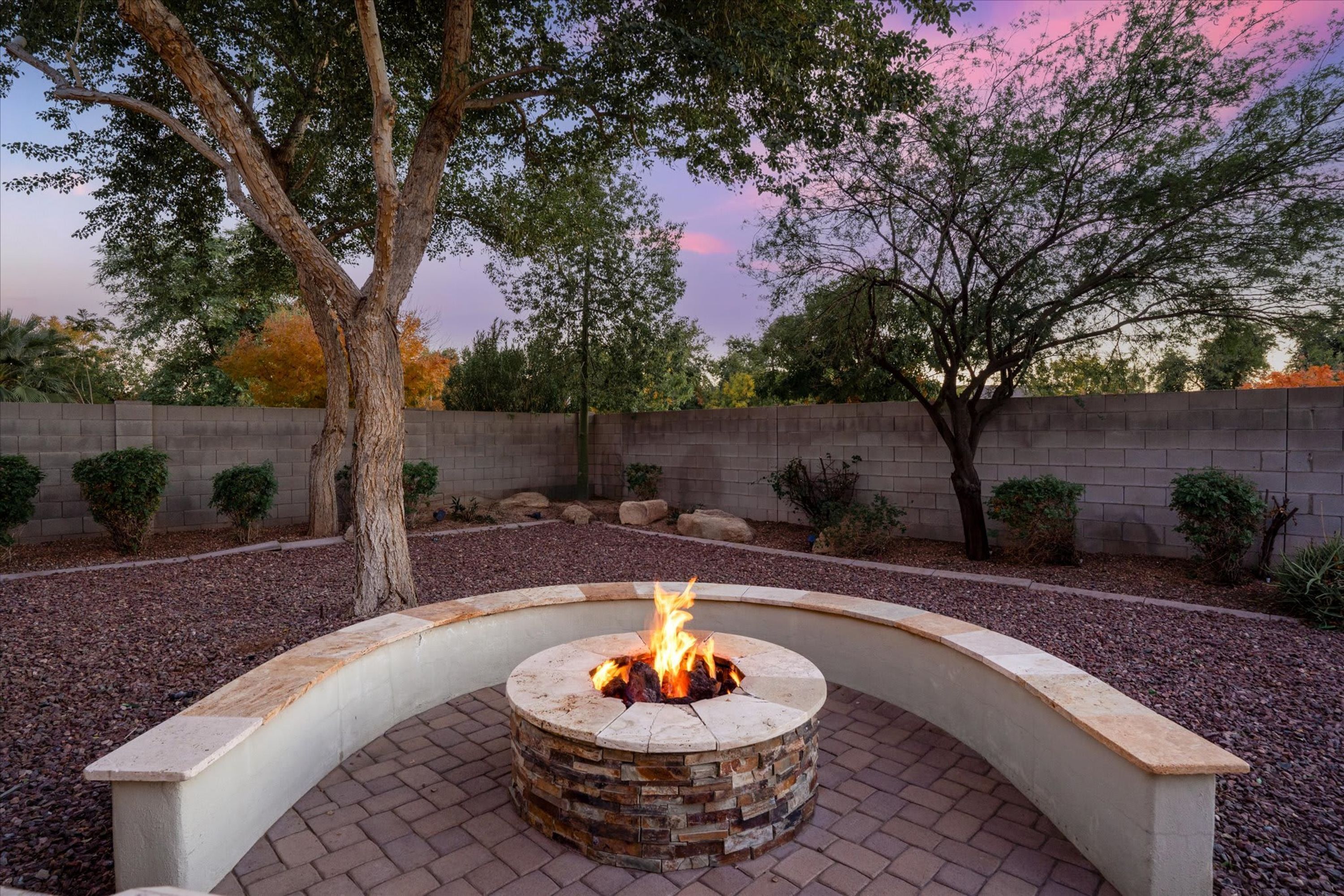 A circular stone fire pit with a fire burning, surrounded by a brick patio and a low stone wall.