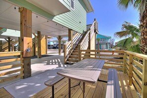 Une terrasse en bois avec une table et des bancs, un espace couvert avec un escalier, et une vue sur un quartier résidentiel avec des maisons et des palmiers.