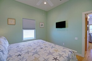 Quarto com cama, janela com persianas, televisão fixada na parede e porta que dá para outro cômodo.