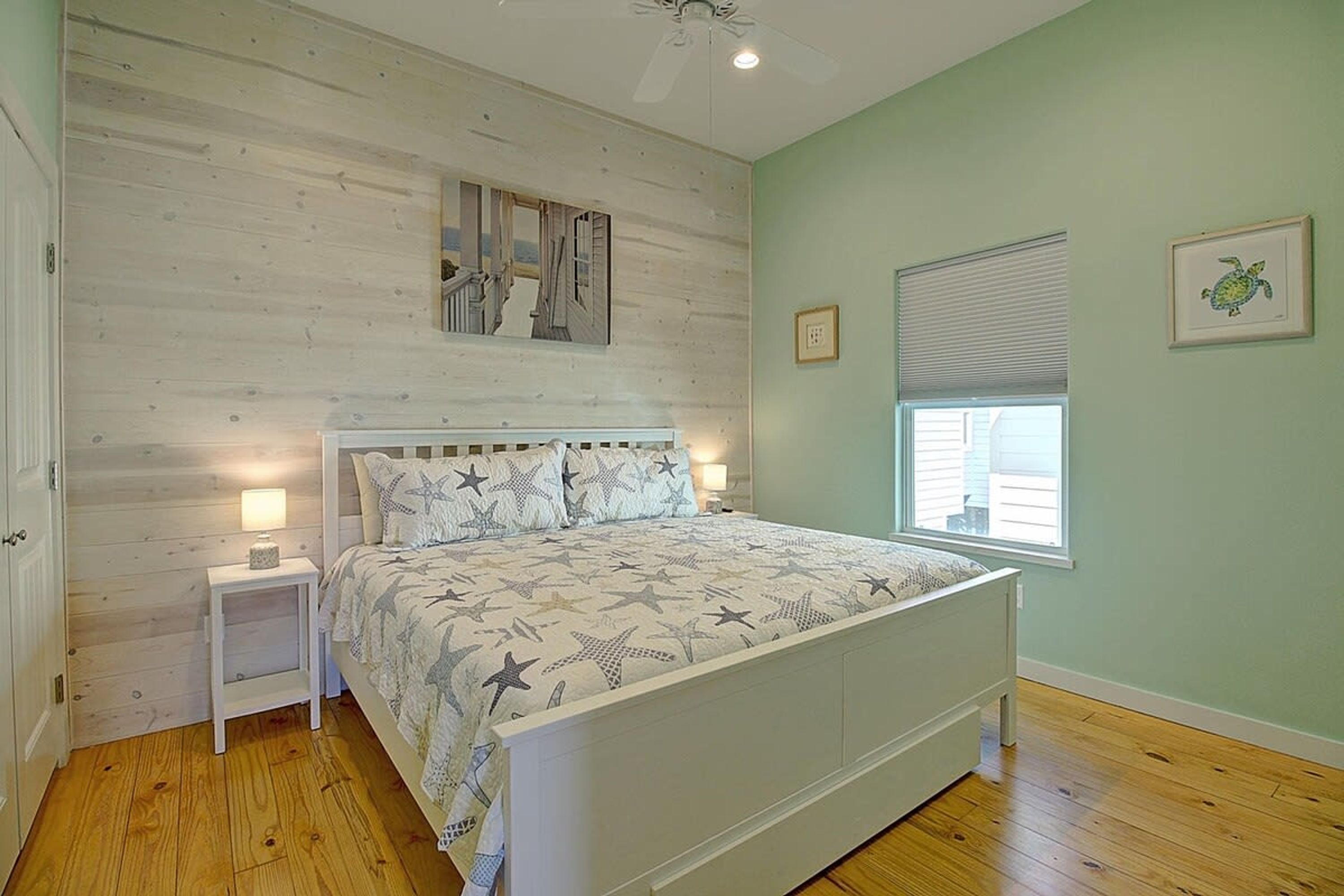 A bedroom with a wooden floor, a bed with a star-patterned bedspread, a nightstand with a lamp, a ceiling fan, and a window with blinds.