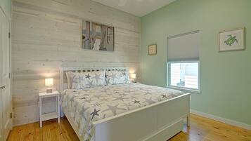 A bedroom with a wooden floor, a bed with a star-patterned bedspread, a nightstand with a lamp, a ceiling fan, and a window with blinds.