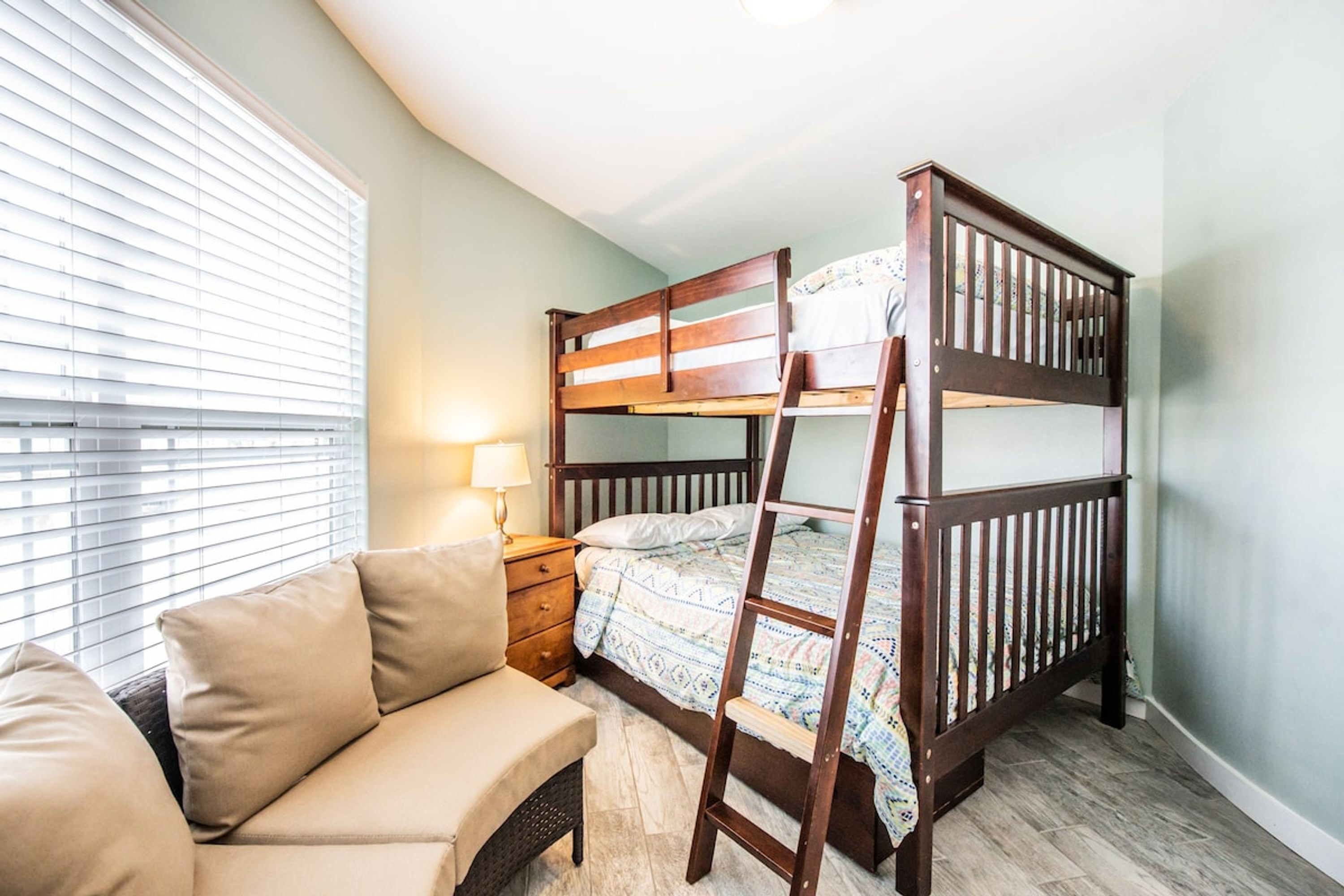 A bunk bed with a ladder, a bedside table with a lamp, and a sofa in a room with light-colored walls and wooden flooring.