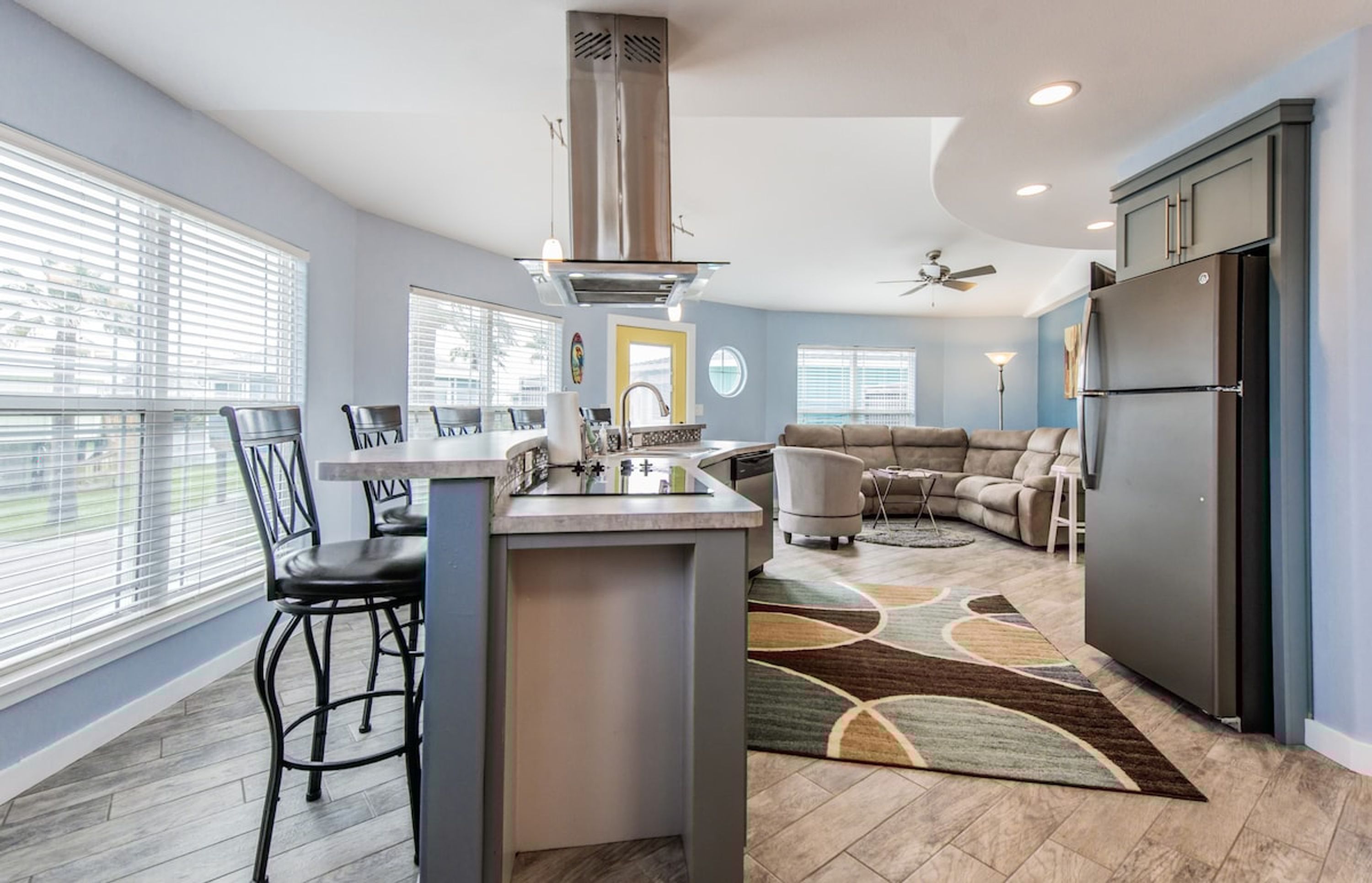 A modern kitchen with a central island, a dining area with chairs, a sofa, and a refrigerator.