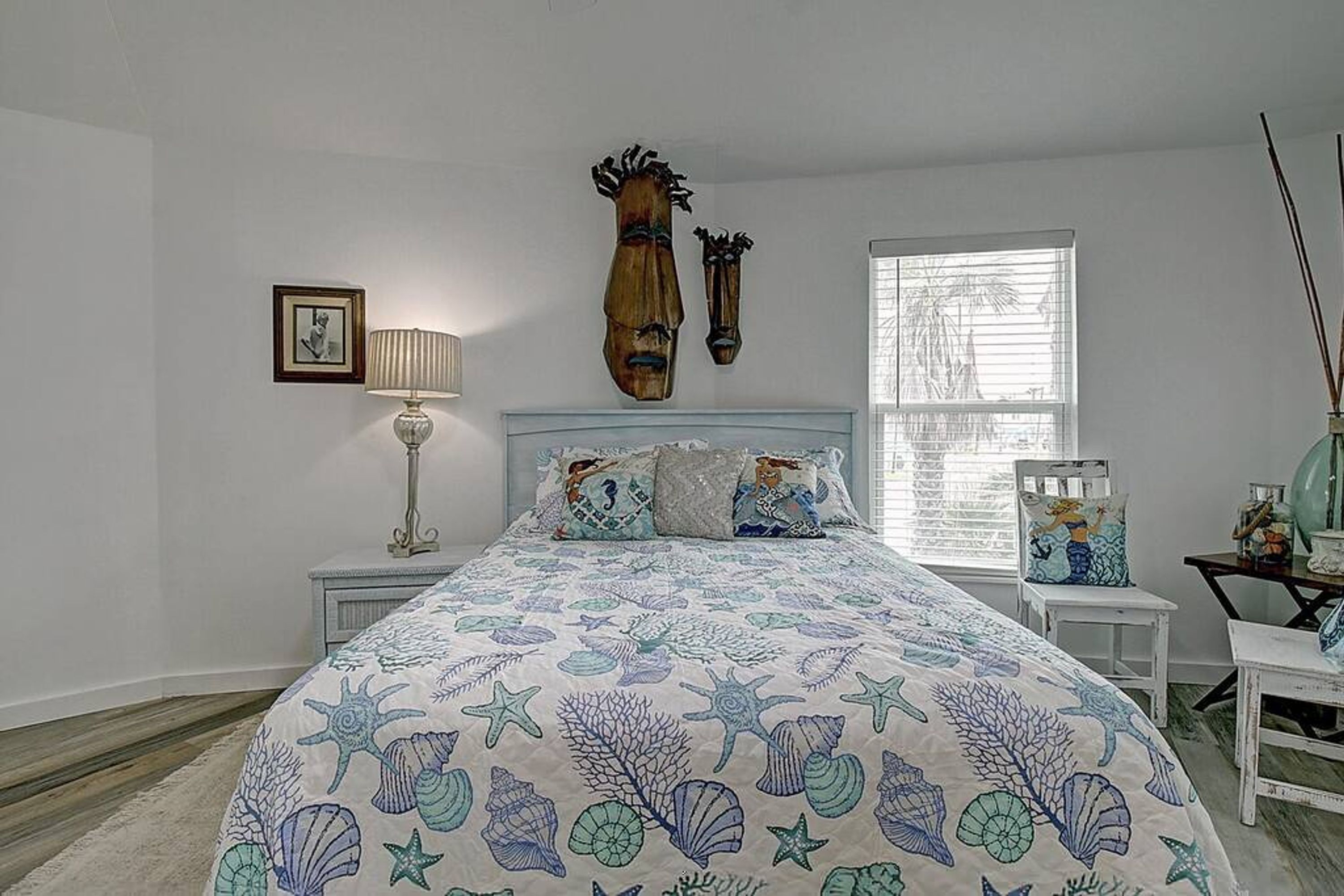 Une chambre à coucher avec un lit, une table de chevet, une lampe, un tableau encadré et une fenêtre avec des stores.