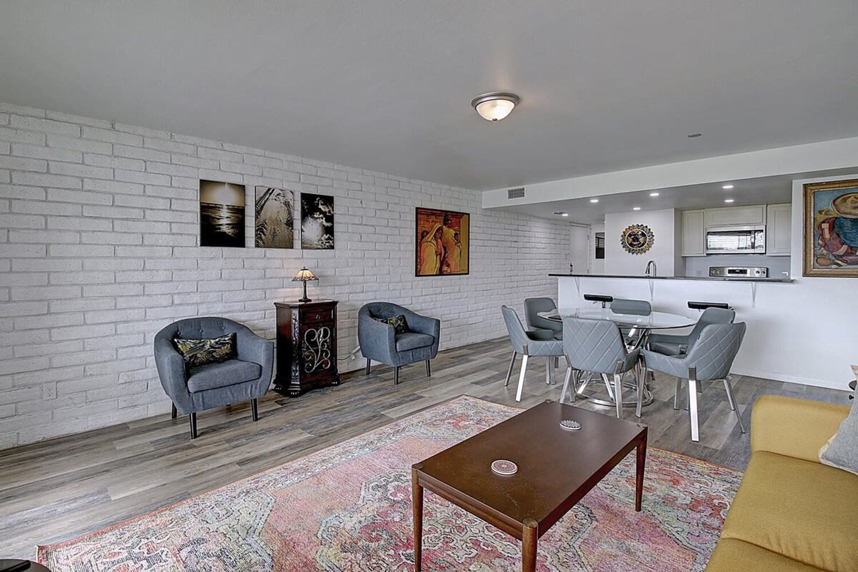 A modern living room with a white brick wall, a dining area with grey chairs, and a kitchen in the background.