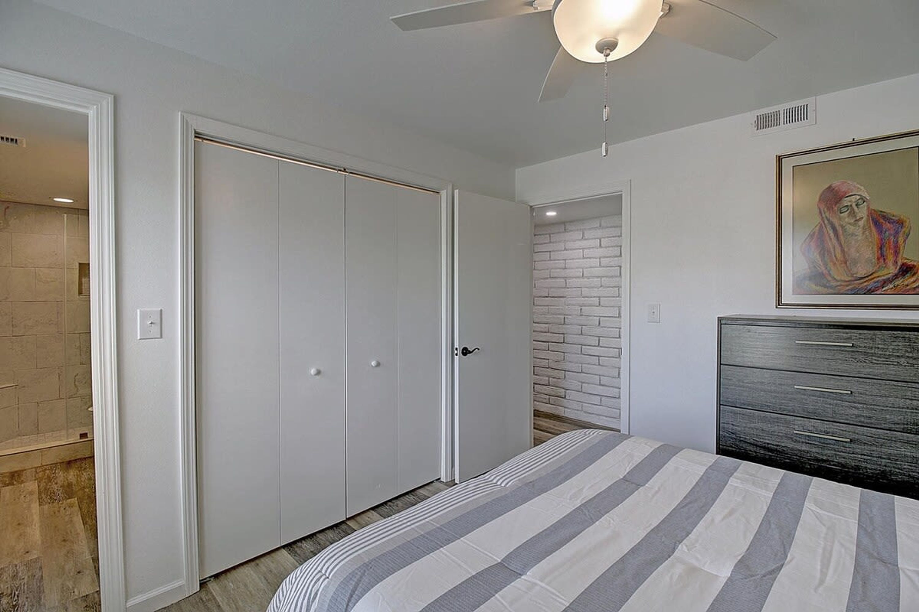 A bedroom with a bed, a dresser, a ceiling fan, and a brick wall.