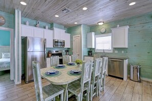 A kitchen with white cabinets, a stainless steel refrigerator, a microwave, and a sink. There is a dining table with chairs and plates. - Blue Wave Community Pool Family Friendly Sleeps 10 (Port Aransas)
