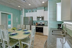 A kitchen with white cabinets, a stainless steel refrigerator, a microwave, and a stove. There is a dining table with chairs and plates set for a meal. - Blue Wave Community Pool Family Friendly Sleeps 10 (Port Aransas)