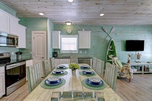 A dining area with a long table set for six, a microwave, and a stove in the kitchen area. - Blue Wave Community Pool Family Friendly Sleeps 10 (Port Aransas)