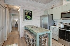 A kitchen with a wooden table, stools, a refrigerator, a microwave, and a stove. - Poco Cabana- Cute Bungalow 2 Community Pools (Port Aransas)