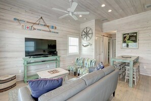 A living room with a television, green furniture, a ceiling fan, and a wall with a compass and a sign. - Poco Cabana- Cute Bungalow 2 Community Pools (Port Aransas)