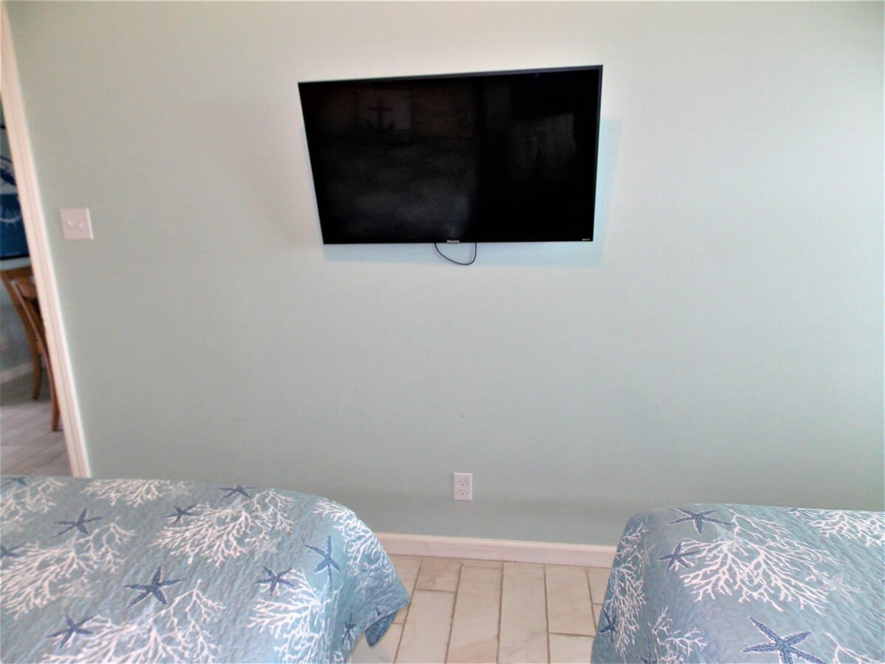 Habitación con televisor de pantalla plana montado en la pared, dos camas con ropa de cama azul y piso de baldosas.